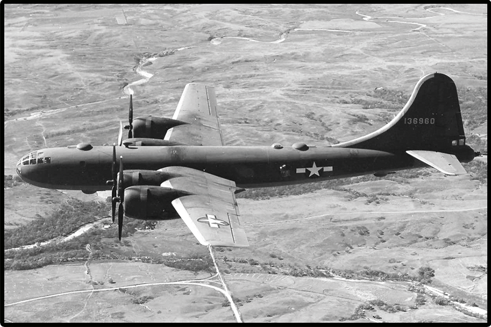 Black and white aerial photograph of a military four-engine propeller plane flying over a textured landscape with roads and fields.
