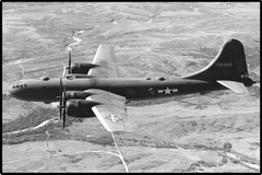 Black and white aerial photograph of a military four-engine propeller plane flying over a textured landscape with roads and fields.
